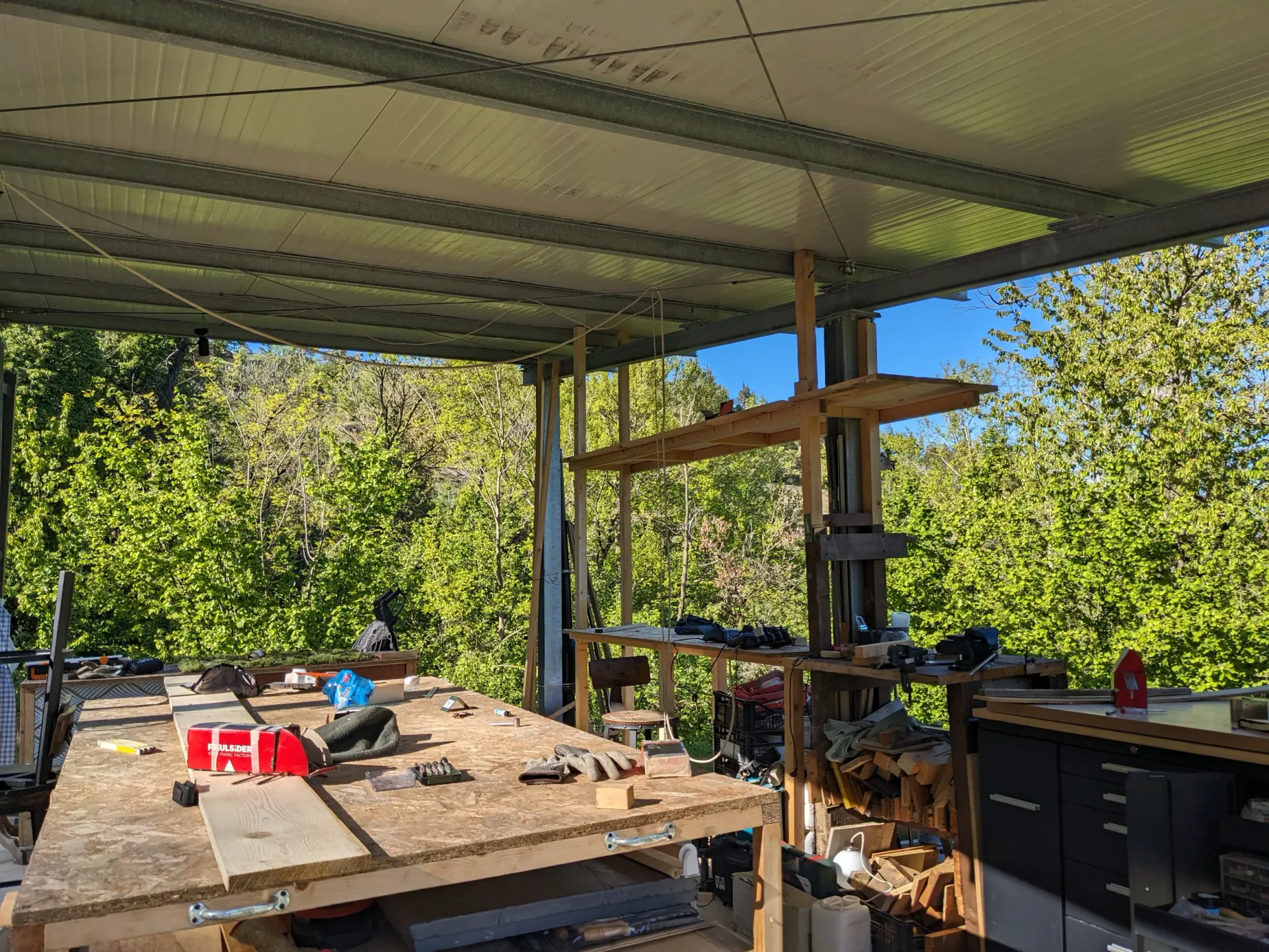 A woodworking workspace under a metal roof with a large wooden workbench in the foreground, tools, gloves, and materials scattered on it, surrounded by shelves, storage units, and lush green trees outside