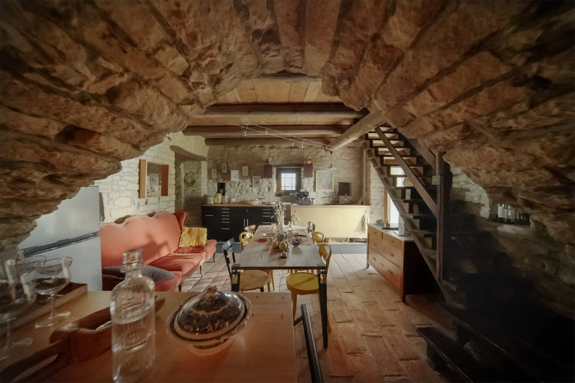 A cozy rustic kitchen/dining area with exposed stone walls and brick ceiling, a wooden staircase to the right, a long dining table with chairs, a red sofa on the left, and various kitchen appliances and decor