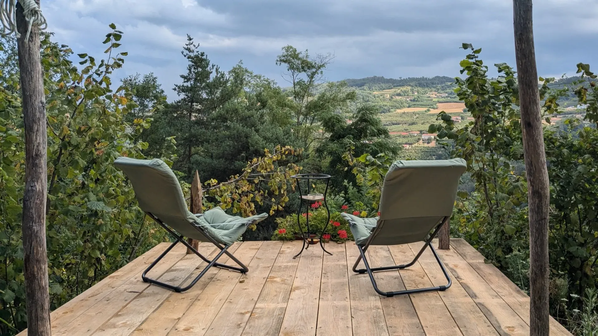 Two chairs on a wooden deck overlooking a lush, green landscape with trees and distant rolling hills under a cloudy sky