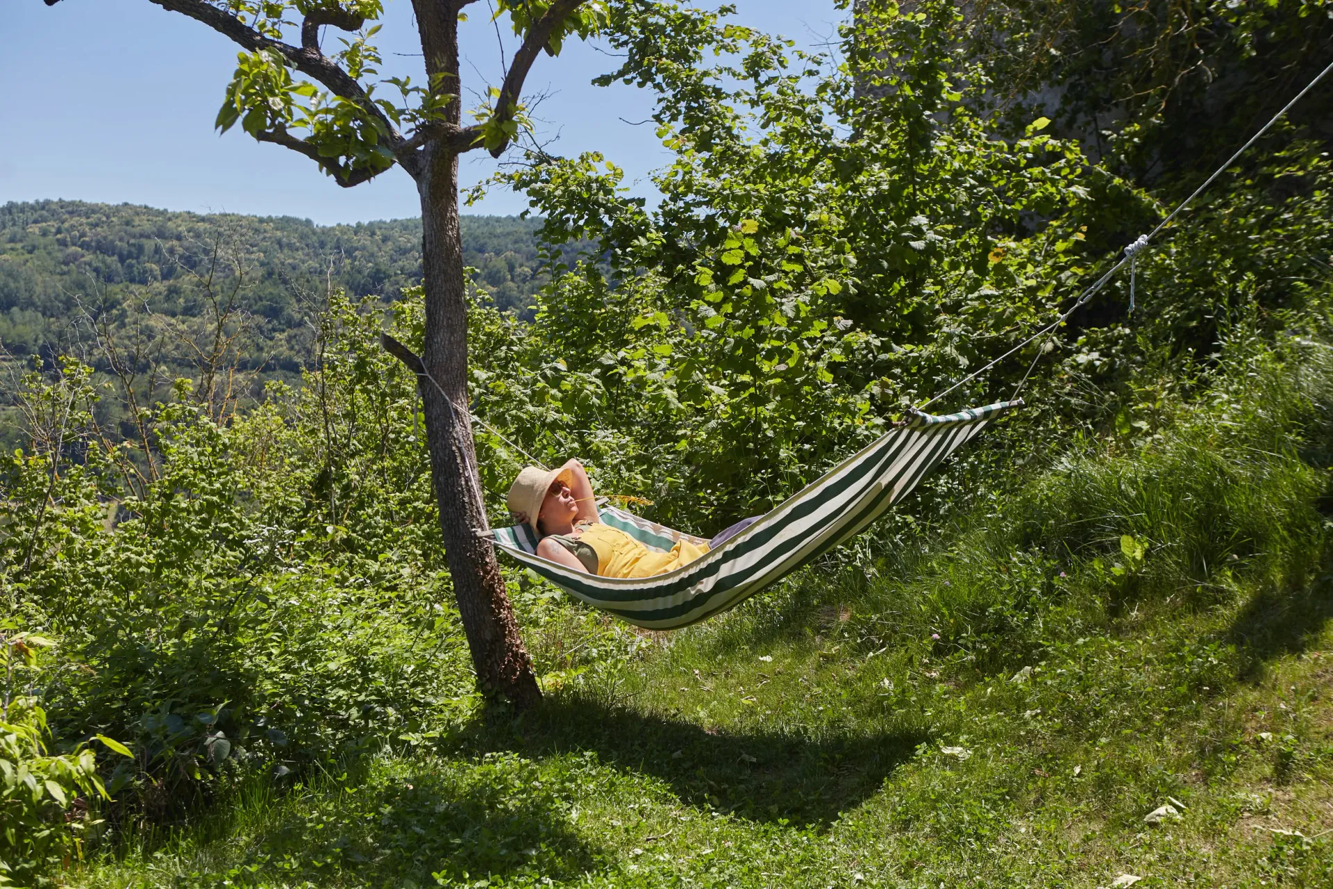 People preparing and cooking pizza outdoors at a picnic table surrounded by trees, with children observing and a little girl in bright pink clothing