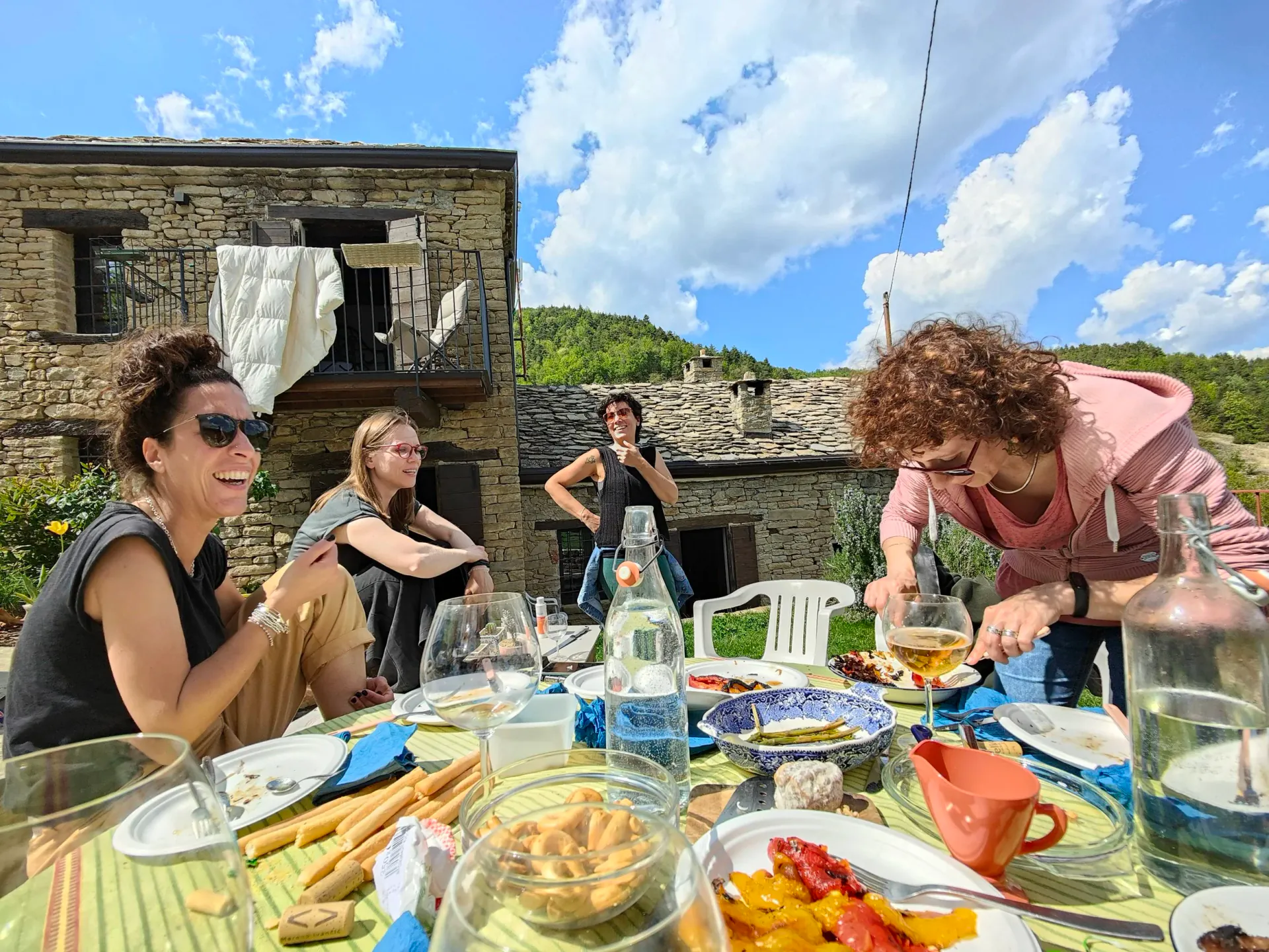 A group of five women enjoying an outdoor meal on a sunny day. They are seated and standing around a table with various dishes and drinks outdoors, with a rustic stone house and green hills in the background