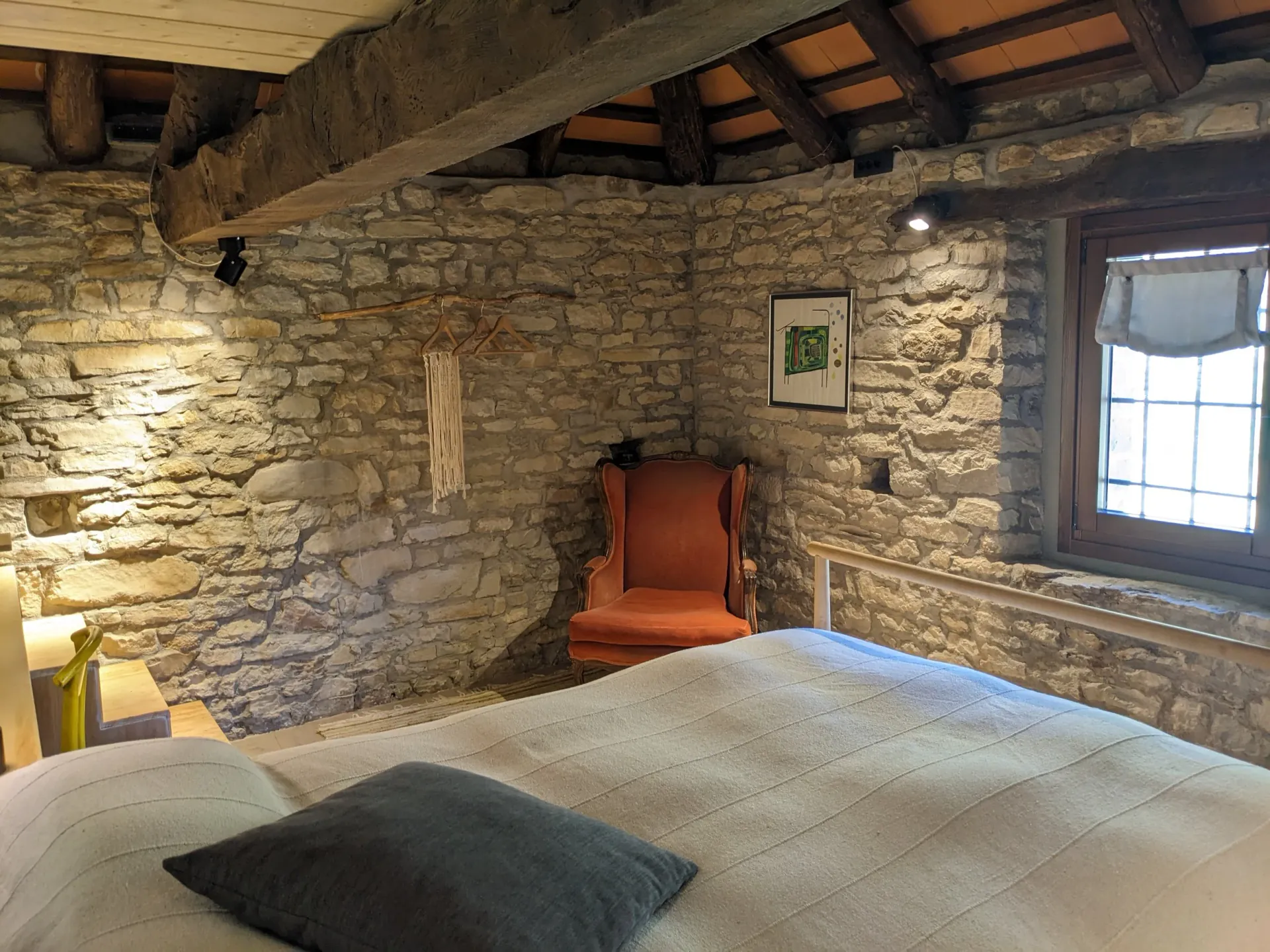 Bedroom with a white bed, a red vintage armchair, stone walls, wooden ceiling beams, and small windows