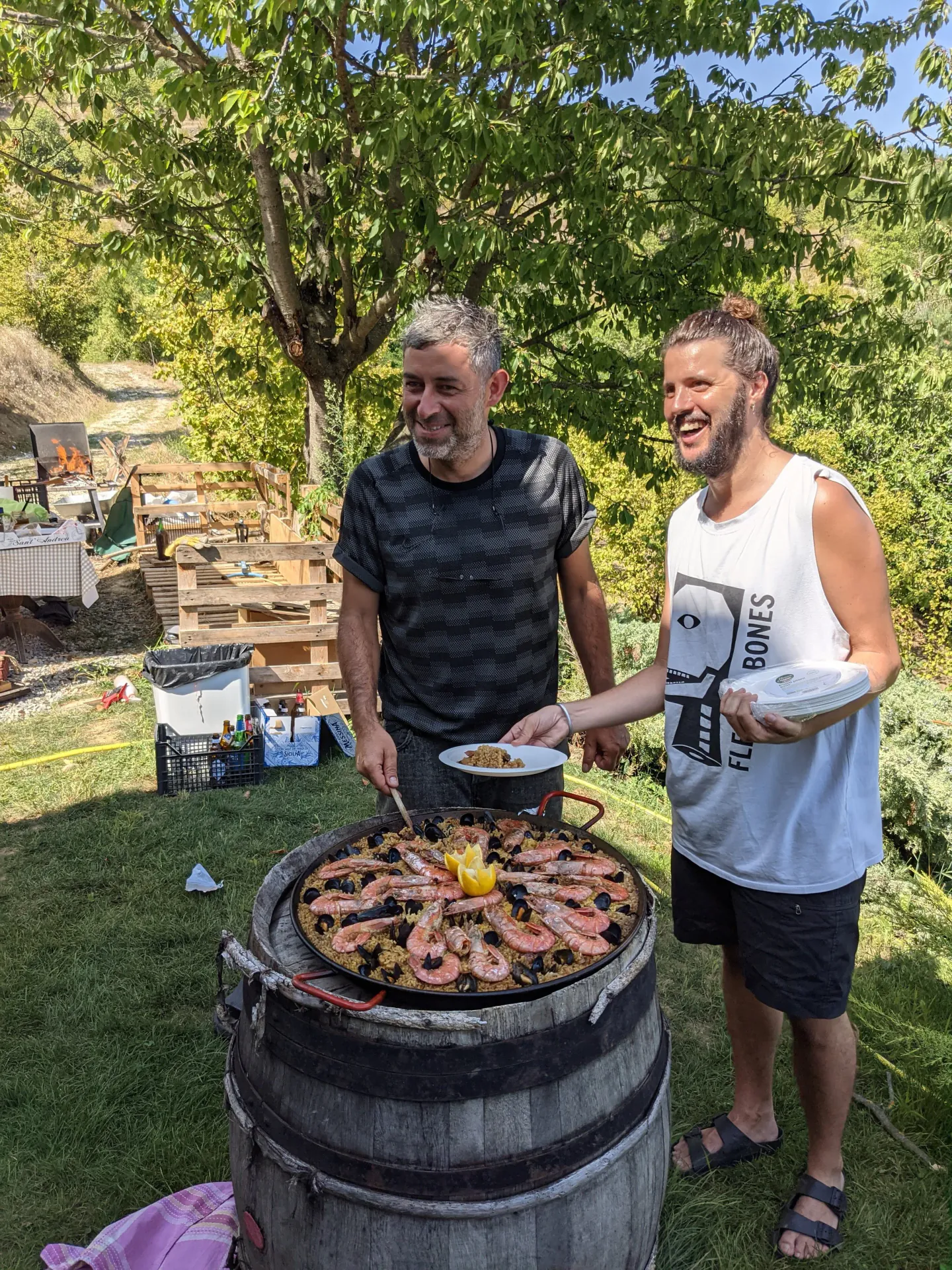 Two men smiling and serving food from a large outdoor black iron skillet with shrimp and lemon slices on top, set on a wooden barrel outside on a sunny day with trees in the background