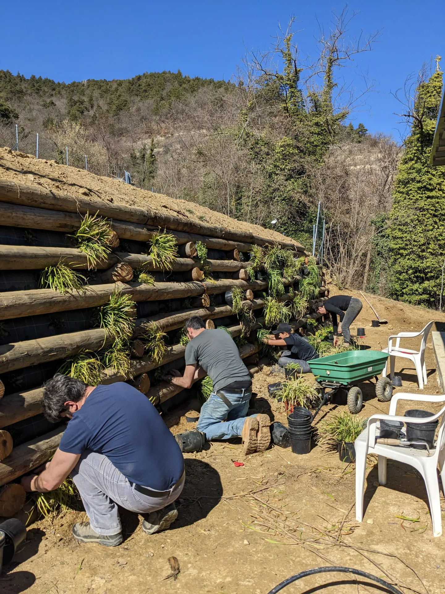 An outdoor patio with a small black metal table, two red metal chairs, a yellow pitcher on the table, and decorative ceramic vases on the ground. A stone wall with steps leading upward, ivy, and hanging baskets are in the background
