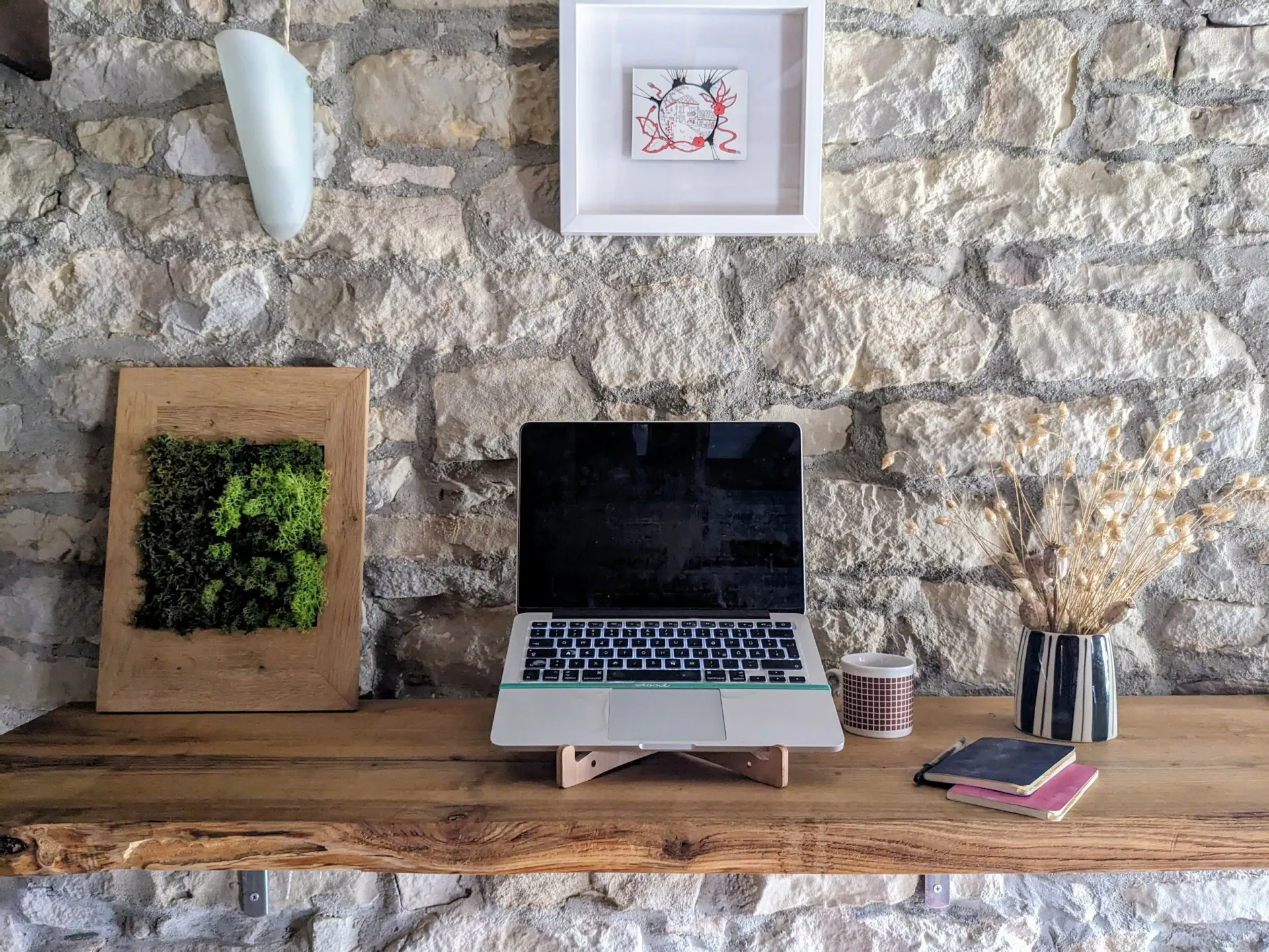 A wooden desk against a stone wall holds an open laptop on a stand, a mug, a small notebook, and a vase with dried flowers. A framed artwork and a decorative wall light are mounted on the stone wall