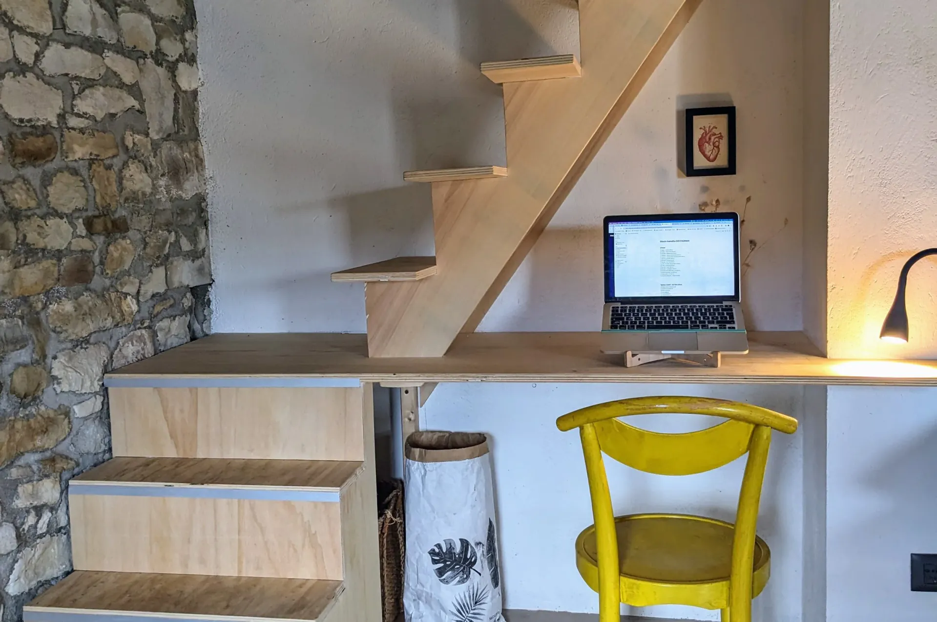 A small workspace with a wooden staircase, a laptop on a wooden desk, a yellow chair, a desk lamp, a framed heart illustration, a stone wall, and a paper bag with floral design