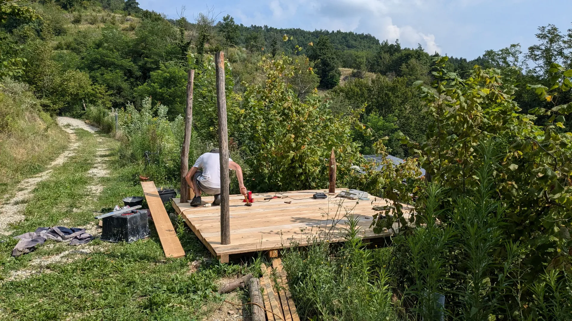A person working on a wooden deck in a rural area with a dirt path and lush green trees and hills in the background
