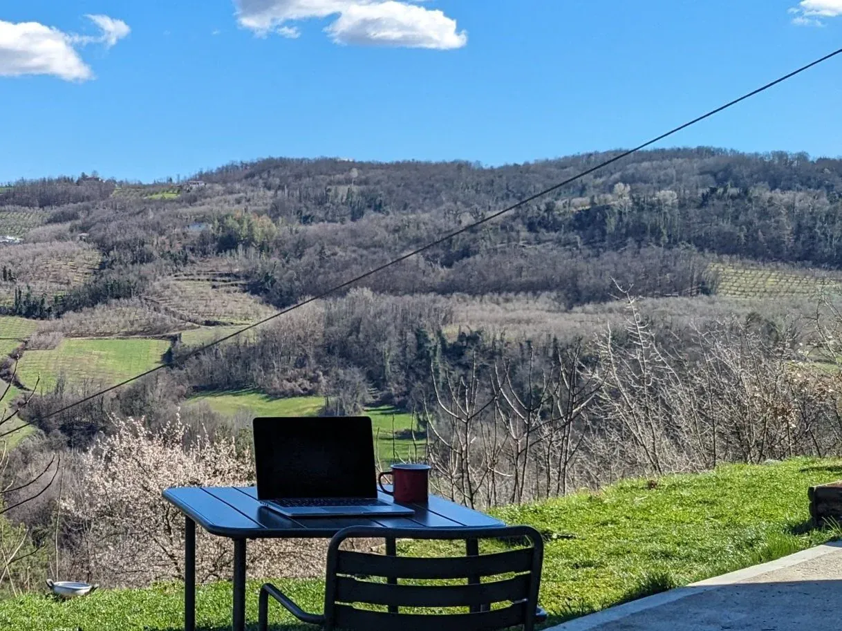 Outdoor scene with a laptop and coffee mug on a table overlooking a hilly landscape with trees and fields in the distance under a partly cloudy sky